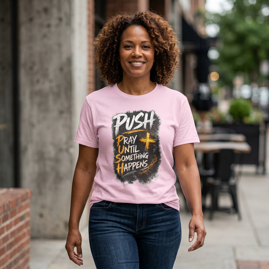 A smiling woman with curly brown hair stands on a city sidewalk wearing a light pink graphic T-shirt and dark blue jeans. The shirt features a distressed black graphic with the words 'PUSH PRAY UNTIL SOMETHING HAPPENS' in white and yellow text.